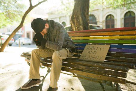 Foto de Deprimido miserable vagabundo sentado en un banco del parque con un cartel de cartón sin trabajo y hambriento - Imagen libre de derechos