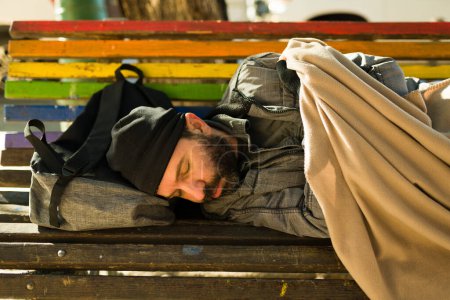 Foto de Hombre solitario sin hogar con una barba durmiendo en el banco del parque cubierto con una manta que parece fría - Imagen libre de derechos