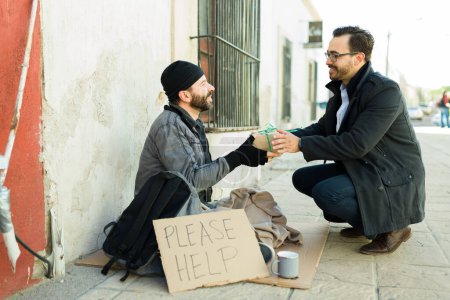 Photo for Cheerful caucasian man smiling while giving food to a hungry happy homeless man asking for help - Royalty Free Image