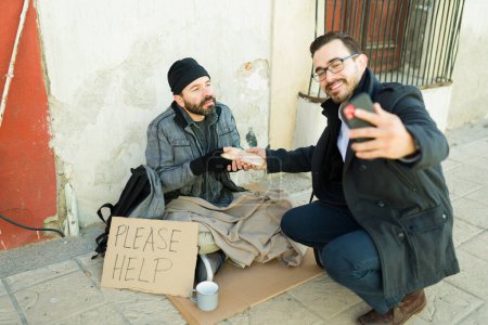 Photo for Hypocrite man doing charity and giving food to a homeless beggar man while taking advantage and taking a selfie - Royalty Free Image