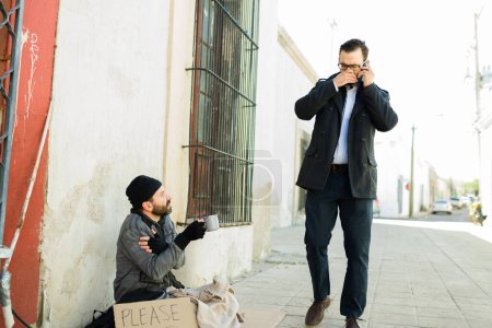 Téléchargez les photos : Homme caucasien couvrant son nez et sa bouche tout en marchant à côté d'un sale sans-abri puant luttant dans la rue - en image libre de droit