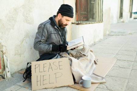 Photo for Homeless man with a beard reading a book while showing a please help cardboard sign while struggling on the street - Royalty Free Image