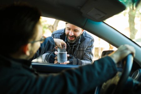 Foto de Feliz hombre sin hogar sonriente recibiendo dinero por comida de un hombre en su coche conduciendo en la calle - Imagen libre de derechos