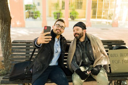 Photo for Happy caucasian man taking a selfie with a homeless man while helping him with charity - Royalty Free Image