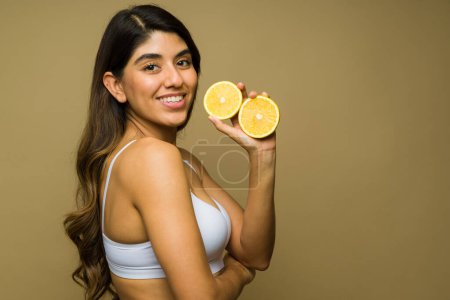 Photo for Profile of a cheerful young woman wearing a white bra taking vitamin c and eating orange fruit for good health and clear skin - Royalty Free Image