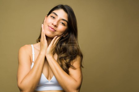 Téléchargez les photos : Latino jeune femme en sous-vêtements toucher son visage et regarder heureux avec sa routine de soins de la peau saine - en image libre de droit