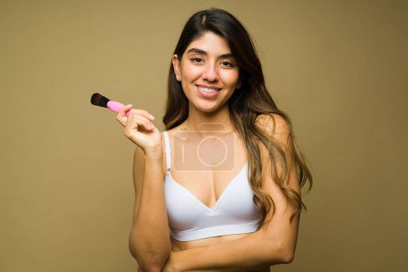 Téléchargez les photos : Jeune femme hispanique en sous-vêtements regardant heureux en utilisant une brosse de maquillage et des produits de beauté tout en se préparant sur un fond de studio - en image libre de droit
