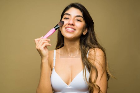 Photo for Smiling latin woman wearing a white bra putting on makeup and using a brush while getting ready with beauty products - Royalty Free Image