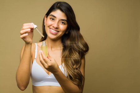 Foto de Emocionada mujer mexicana sonriendo usando productos para el cuidado de la piel y sosteniendo una botella de suero de ácido hialurónico - Imagen libre de derechos