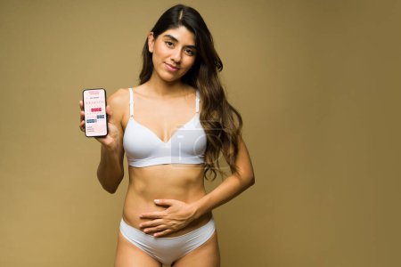 Photo for Sad beautiful hispanic woman suffering from pain cramps and showing a period app and calendar on her smartphone - Royalty Free Image