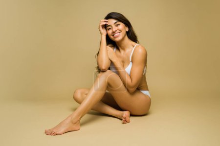 Preciosa joven latina sonriendo en ropa interior para un concepto de belleza mientras se relaja contra un fondo de estudio