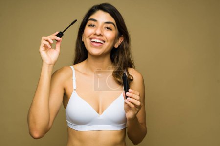 Foto de Mujer joven emocionada riendo mientras se pone rímel y productos de maquillaje contra un fondo de estudio - Imagen libre de derechos