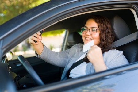 Foto de Mujer gorda feliz tomando una selfie mostrando su nueva licencia de conducir con su teléfono inteligente mientras conduce su coche - Imagen libre de derechos