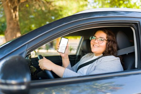 Photo for Attractive happy fat woman smiling working as a driver showing her smartphone using a ride share app while driving a car - Royalty Free Image