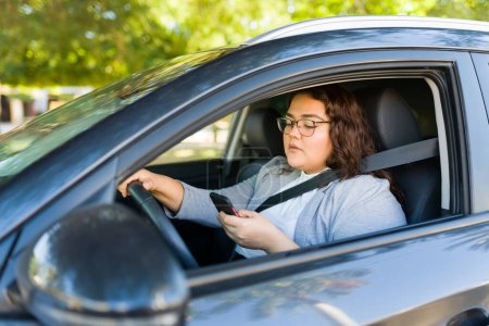 Foto de Mujer latina obesa ocupada conduciendo y enviando mensajes de texto en su teléfono inteligente o trabajando como conductora mientras usa una aplicación para compartir viajes - Imagen libre de derechos