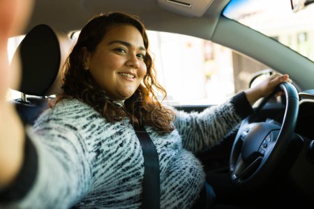Photo for Personal perspective of a fat latin woman looking happy and taking a selfie while driving her car - Royalty Free Image
