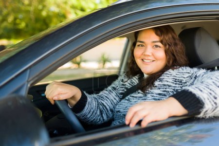 Téléchargez les photos : Attrayant gai grosse femme souriante regardant la caméra tout en conduisant sa voiture et en voyageant - en image libre de droit
