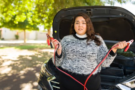 Foto de Mujer gorda frustrada haciendo contacto visual sosteniendo los cables de salto de su coche roto sin batería - Imagen libre de derechos