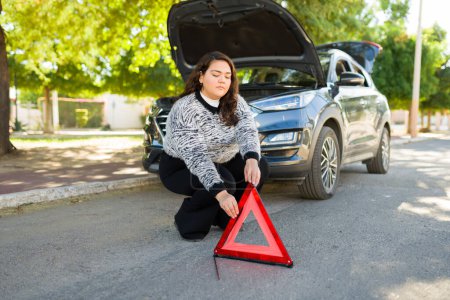 Photo for Upset fat woman using a warning triangle to prevent accidents because of her broken down car - Royalty Free Image