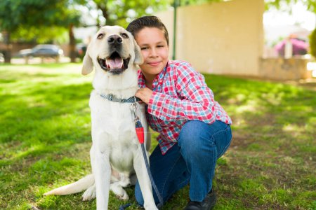 Téléchargez les photos : Portrait d'un adorable garçon latino souriant et embrassant son copain labrador chien tout en jouant ensemble dans le parc - en image libre de droit