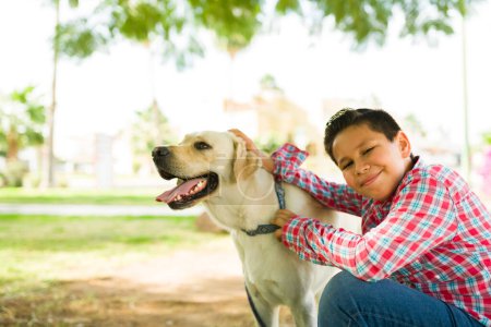 Téléchargez les photos : Adorable enfant de 8 ans souriant tout en caressant et en donnant de l'amour à son chien labrador heureux tout en jouant à l'extérieur - en image libre de droit