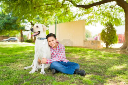 Foto de Alegre niño hispano abrazando a su perro recuperador feliz y sonriendo mientras hace contacto visual y se divierte con su amigo en el parque - Imagen libre de derechos