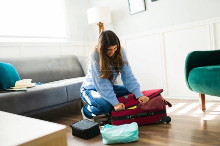 Foto de Mujer latina molesta preparándose para viajar y tratando de cerrar su maleta llena de ropa y luciendo estresada antes de su viaje - Imagen libre de derechos