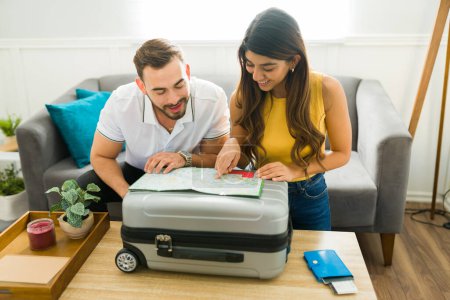 Photo for Cheerful young woman and man looking at a map and preparing a suitcase while planning a last minute trip together - Royalty Free Image