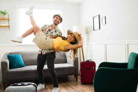 Téléchargez les photos : Heureux homme caucasien portant sa petite amie et riant à la maison tout en étant prêt pour leurs vacances d'été - en image libre de droit