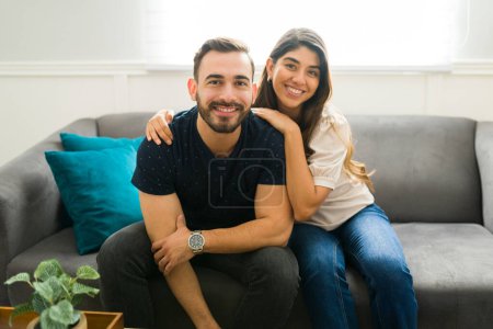 Photo for Beautiful young couple in love hugging and looking at the camera while relaxing in the living room at home - Royalty Free Image