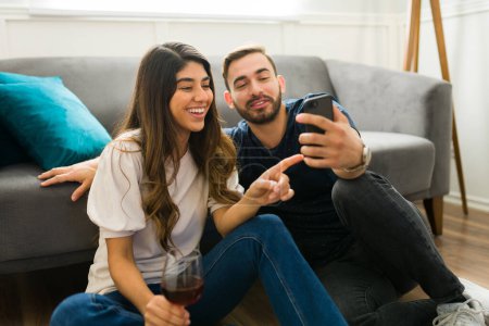Téléchargez les photos : Jeune couple excité regardant les médias sociaux sur le smartphone tout en buvant du vin ensemble à la maison - en image libre de droit
