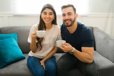 Photo for Cheerful beautiful couple drinking coffee at home and smiling while making eye contact - Royalty Free Image