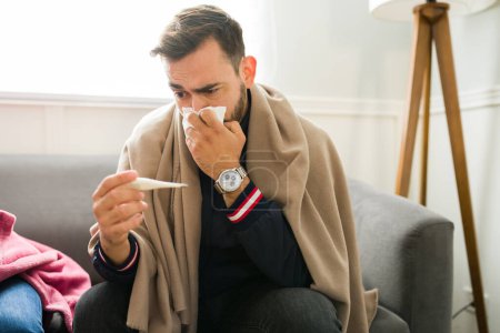 Téléchargez les photos : Malade homme caucasien avec une fièvre à l'aide du thermomètre et se moucher souffrant d'un rhume - en image libre de droit