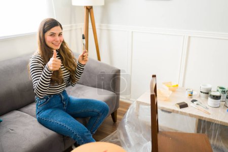 Foto de Mujer joven emocionada dando un pulgar hacia arriba antes de comenzar un proyecto de volteo de muebles en casa - Imagen libre de derechos