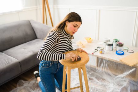 Téléchargez les photos : Belle femme utilisant du papier abrasif sur un mobilier de tabouret et travaillant sur un projet d'amélioration de la maison - en image libre de droit