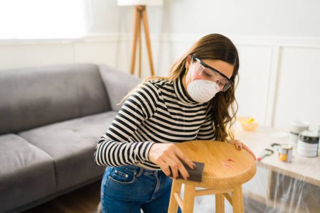 Foto de Mujer joven con gafas de seguridad y mascarilla usando papel de lijar en un mueble de taburete - Imagen libre de derechos