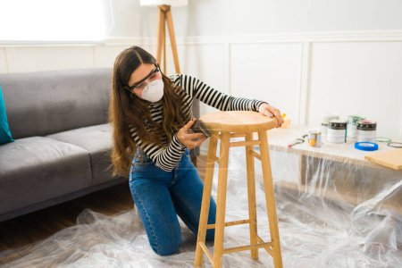 Téléchargez les photos : Belle femme portant des lunettes de sécurité et un masque facial se préparant à peindre un tabouret tout en faisant des meubles flipping - en image libre de droit