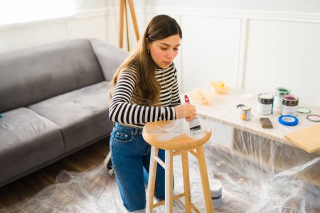 Téléchargez les photos : Belle jeune femme peignant un tabouret sur son bricolage projet d'amélioration de la maison ou de meubles retournant - en image libre de droit