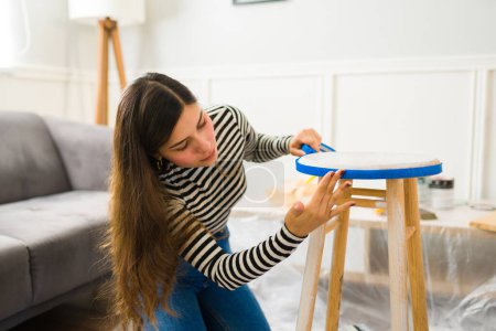 Foto de Hermosa mujer con un negocio de restauración de muebles poniendo cinta adhesiva en un taburete para empezar a pintar - Imagen libre de derechos