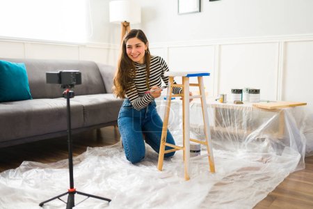 Happy woman smiling filming a video with her smartphone while doing furniture flipping work and painting a stool