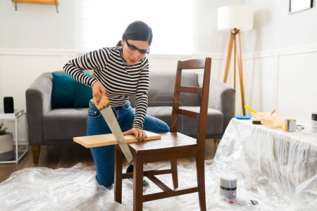 Foto de Mujer joven trabajando en su proyecto de negocio de restauración de muebles volteo mientras corta madera - Imagen libre de derechos