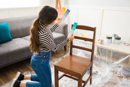Téléchargez les photos : Belle femme faisant un projet de restauration de meubles bricolage et regardant des échantillons de couleur pour peindre une chaise - en image libre de droit