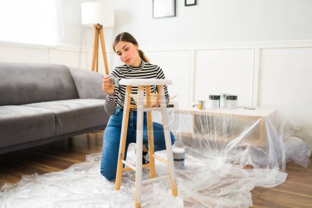 Téléchargez les photos : Belle femme travaillant sur son entreprise de restauration de meubles tout en peignant une chaise de tabouret - en image libre de droit
