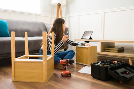 Téléchargez les photos : Jeune femme regardant un tutoriel vidéo sur la tablette pour des instructions pour assembler des meubles à la maison - en image libre de droit