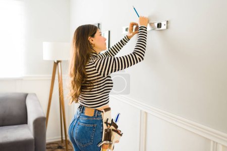 Photo for Hispanic woman using her tools and a level while hanging decorations or pictures on the wall - Royalty Free Image