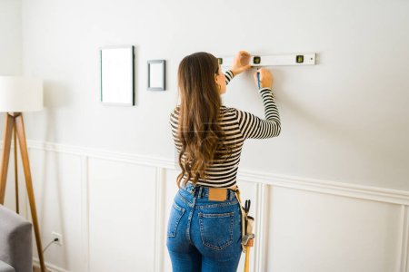 Foto de Hermosa mujer vista desde atrás usando una herramienta de nivel y un martillo para colgar decoraciones de imágenes en la pared de su sala de estar - Imagen libre de derechos