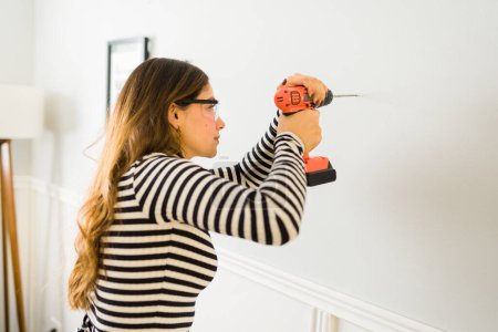 Téléchargez les photos : Femme latine avec des lunettes de sécurité en utilisant une perceuse sur le mur pour accrocher des photos ou des décorations - en image libre de droit