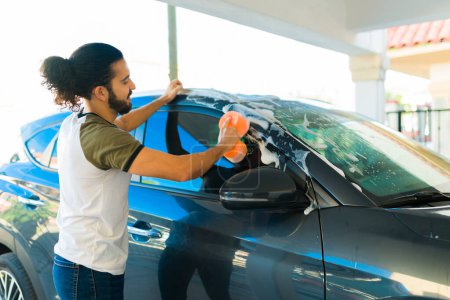 Photo for Happy latin man seen from behind washing his car with a sponge and soap while doing chores in his home garage - Royalty Free Image