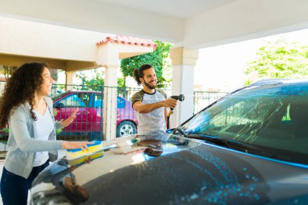 Foto de Guapo feliz hombre usando una manguera para lavar su coche con jabón junto con su novia - Imagen libre de derechos