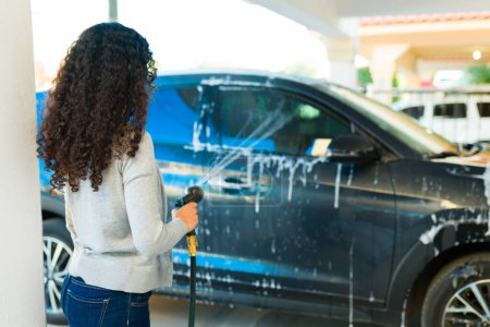 Téléchargez les photos : Femme latine avec les cheveux bouclés à l'extérieur en utilisant un tuyau d'eau à la maison pour laver sa voiture avec du savon - en image libre de droit